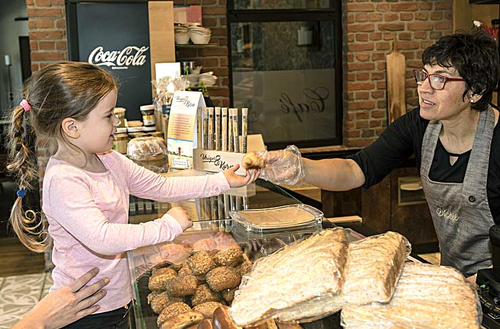 Symboldbild für Einkaufen: Eine Bäckertheke mit Brötchen und Kuchen, die Verkäuferin reicht einem kleinen Mädchen etwas an
