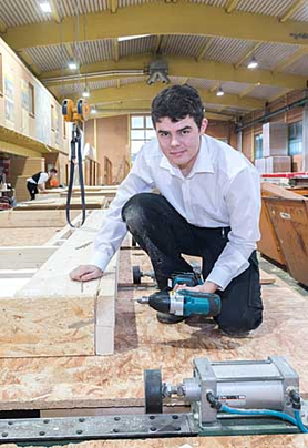 Der Azubi einer Zimmerei kniet auf Spanplatten und hält eine Bohrmaschine in der Hand