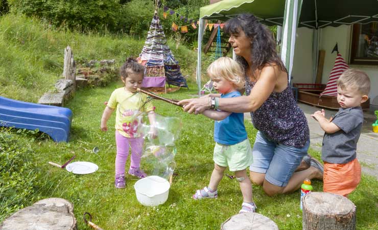 Betreuerin mit drei Kleinkindern im Garten, es werden gerade Rieseseifenblasen produziert