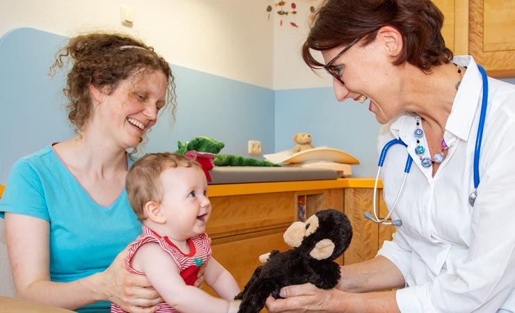 In der Kinderarztpraxis: Mutter mit Kleinkind und Kinderärztin sitzen sich gegenüber, die Ärztin gibt dem Kleinkind ein Kuscheltier