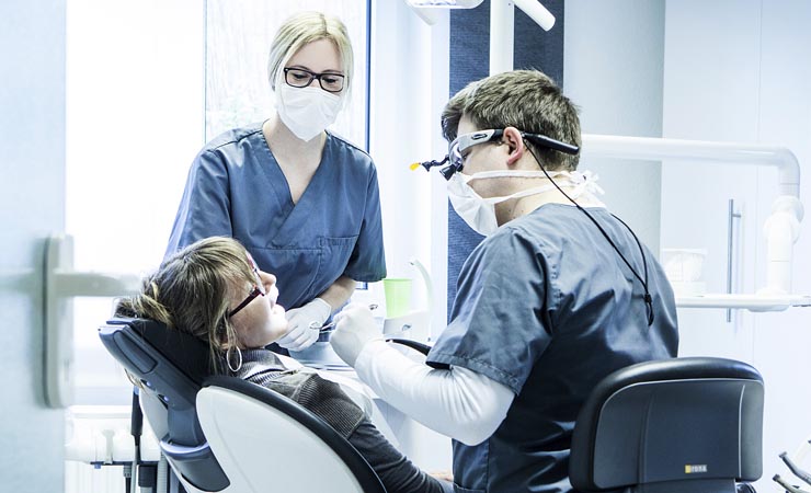 In der Zahnarztpraxis: Eine Patientin im Behandlungsstuhl öffent den Mund, der Zahnarzt und die Helferin behandeln