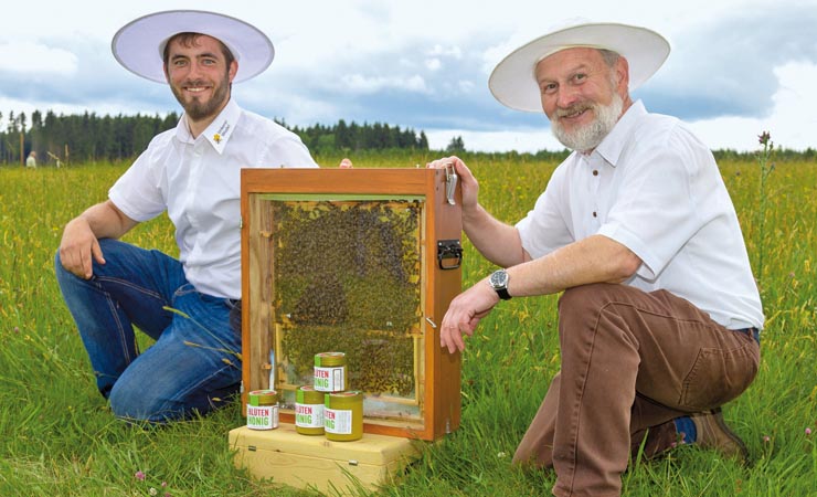 Zwei Imker mit Hüten auf einer Wiese, dazwischen eine Bienenwabe und Honiggläser